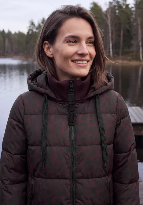 Een glimlachende vrouw in een doorgestikte jas staat aan de oever van een meer met bos op de achtergrond.