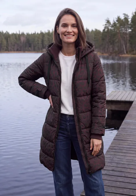 Een glimlachende vrouw staat op een steiger bij een meer en draagt een lange winterjas.