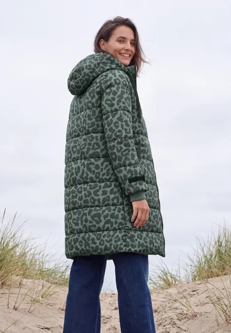 Een glimlachende vrouw in een groen gewatteerde jas met capuchon staat op zandduinen bij het strand.