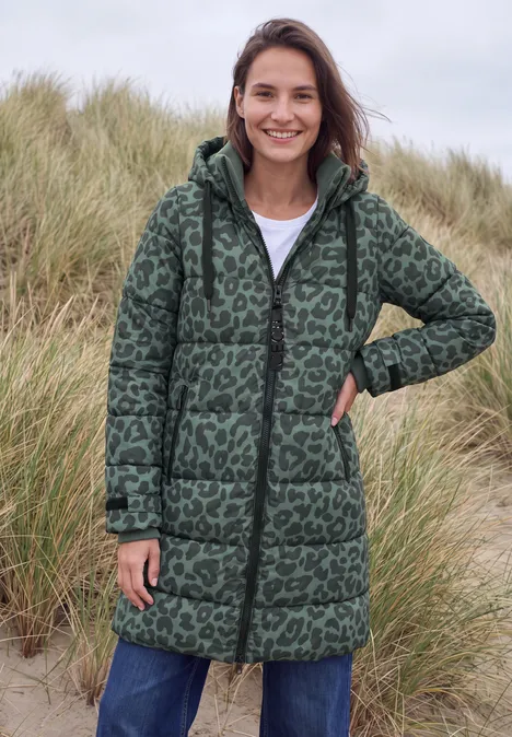 Vrouw in groene gewatteerde jas met luipaardprint, glimlachend voor duinen.