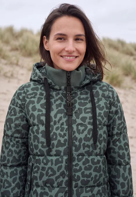 Jonge vrouw in groene gewatteerde jas met dierenprint, glimlachend voor duinen aan het strand.