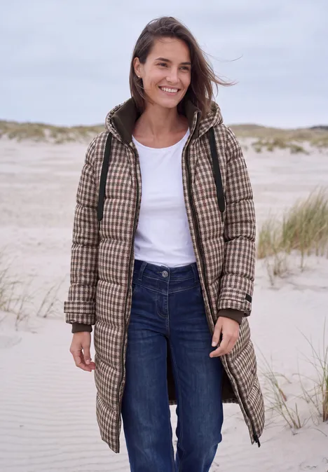 Vrouw lacht op het strand in een lange geruite jas, wit shirt en spijkerbroek.