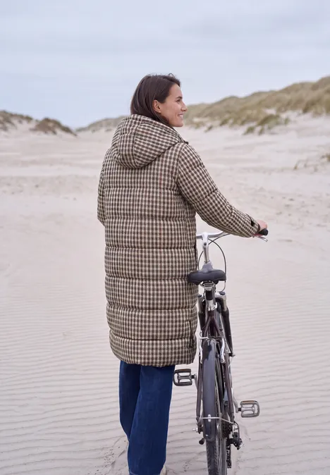 Vrouw duwt haar fiets over het strand in een lange geruite jas, met duinen op de achtergrond.