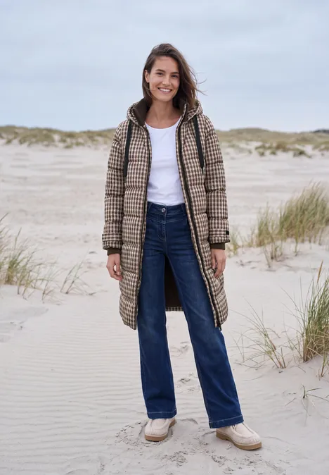 Een lachende vrouw in geruite jas, jeans en instappers staat op een winderig zandstrand tussen duinen.