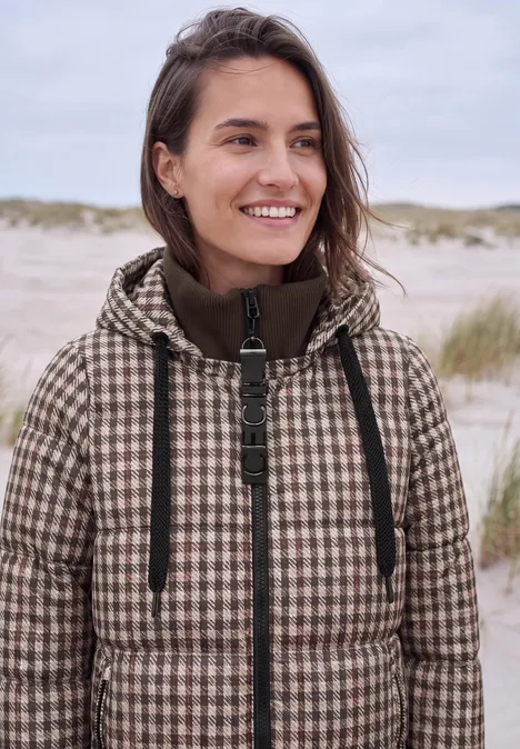 Een glimlachende vrouw in een pied-de-poule jas op het strand voor duinen.