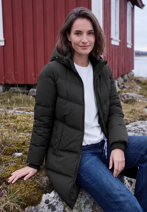Frau in schwarzer Winterjacke sitzt vor rotem Holzhaus an Küste, trägt weiße Bluse und Jeans.