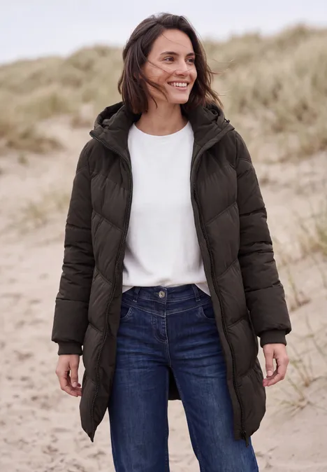 Frau in langer dunkelgrüner Steppjacke, weißem Shirt und Jeans, lächelt vor Dünenhintergrund.