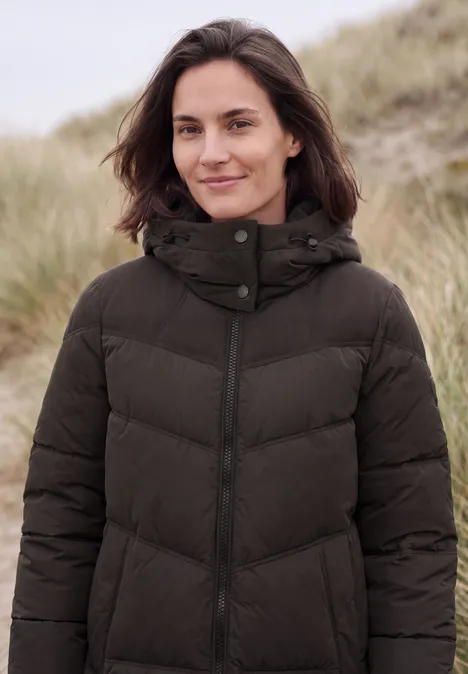 Eine Frau in dunkler Steppjacke steht lächelnd in Dünenlandschaft.