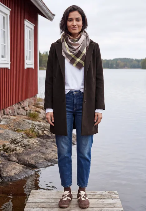 Eine Frau im Mantel, Jeans und Schal steht auf einem Steg vor einem roten Bootshaus am See.