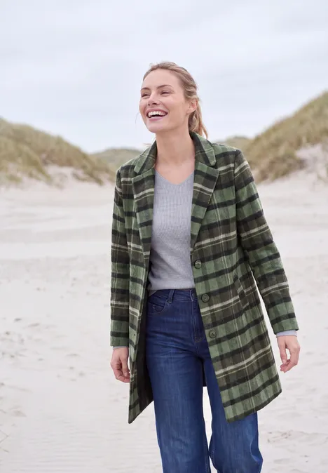 Reversmantel in Wolloptik shadow khaki Eine lächelnde Frau in grün kariertem Mantel, grauem Shirt und Jeans am Strand vor Dünen.