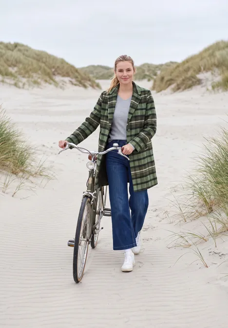 Reversmantel in Wolloptik shadow khaki Frau schiebt Fahrrad über Dünenstrand, trägt grün karierten Mantel, Jeans und weiße Sneakers.