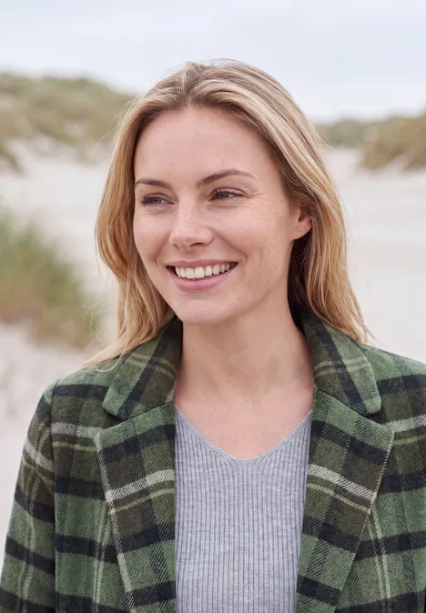 Reversmantel in Wolloptik shadow khaki Eine lächelnde Frau in karierter grüner Jacke steht am Strand vor Dünen.