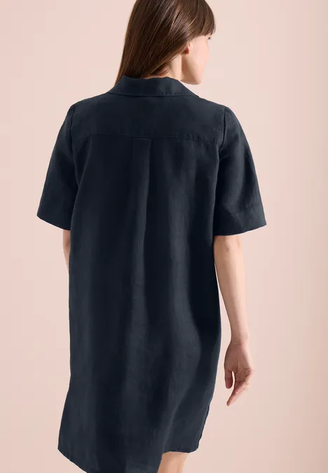 Rückenansicht einer Frau im kurzärmeligen, knielangen, dunklen Hemdkleid vor rosafarbenem Hintergrund.