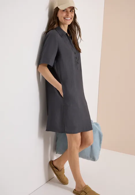 Frau lehnt in einem dunkelgrauen Kurzarm-Hemdkleid und geflochtenen Keilsandalen an einer Wand.