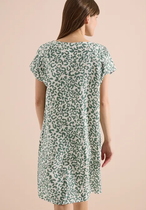 Rückansicht einer Frau in locker sitzendem, grün-weißem Leopardenkleid vor hellem Hintergrund.