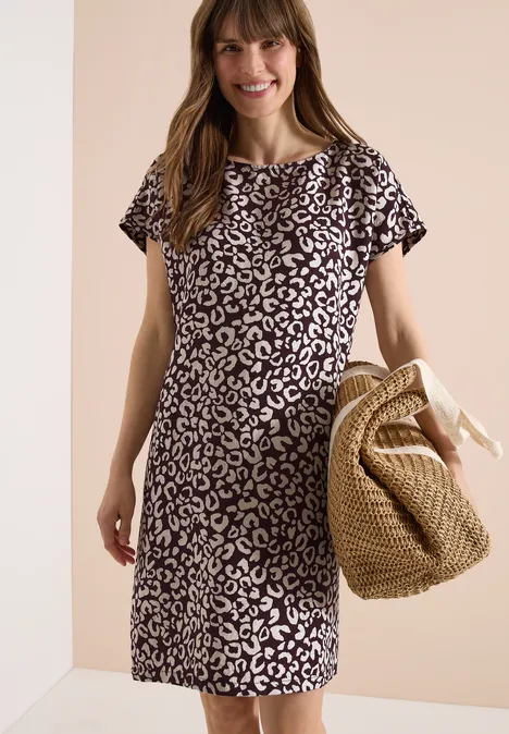 Frau trägt ein kurzärmliges, knielanges schwarz-weißes Leopardenkleid und hält eine geflochtene Strohtasche.