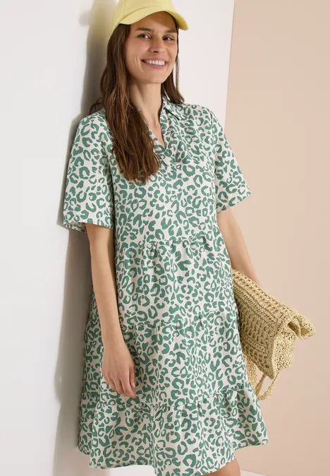Femme appuyée sur un mur, robe chemise ample à manches courtes avec motif léopard vert, sac en paille.