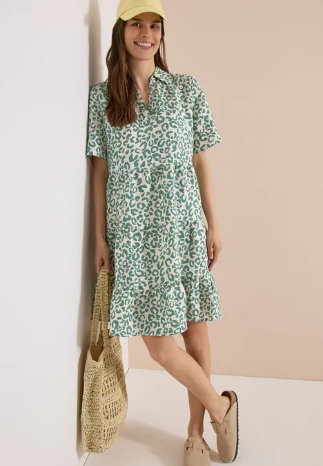 Femme en robe d'été verte à fleurs blanches, manches courtes et volants, avec sac en paille et espadrilles.