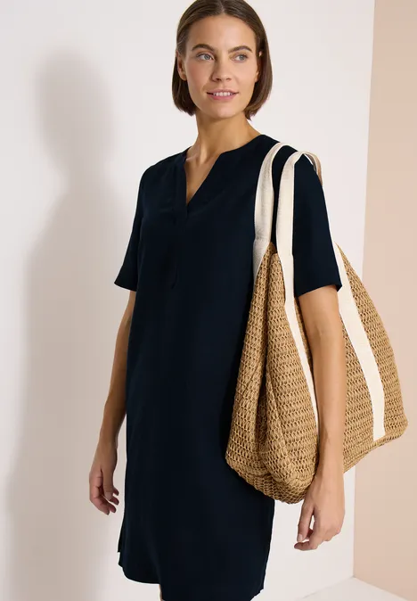 Frau in dunkelblauem knielangem V-Ausschnitt-Kleid mit kurzen Ärmeln und großer Strohtasche an der Schulter.
