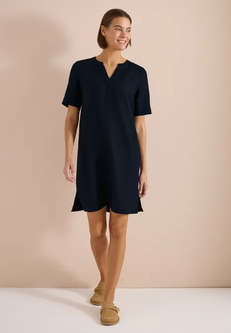 Frau in schlichtem dunkelblauem Etuikleid und beigen Slippers vor neutralem Hintergrund.