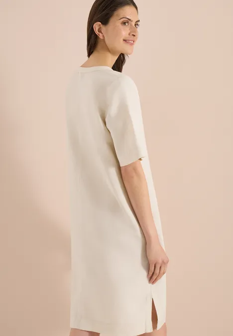 Rückenansicht einer Frau in einem schlichten, cremefarbenen Midi-Kleid mit kurzen Ärmeln und seitlichem Schlitz.