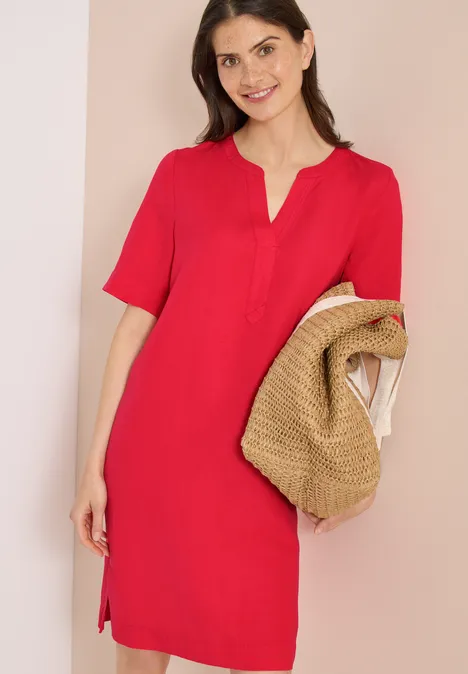 Eine Frau trägt ein rotes, knielanges Kleid mit V-Ausschnitt und hält einen Strohhut.