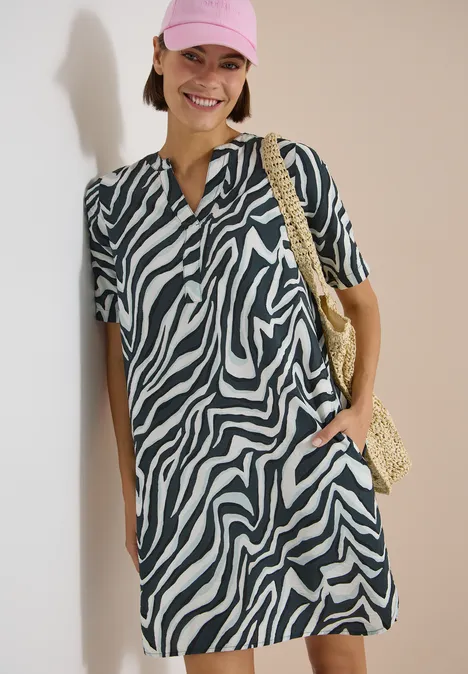 Frau trägt kurzärmeliges Zebra-Kleid und Basttasche vor hellem Hintergrund.