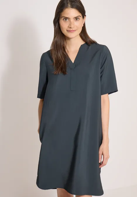 Knie-lange jurk met split in de hals palm breeze green Een vrouw draagt een losvallende, knielange, donkergroene jurk met korte mouwen voor een neutrale achtergrond.