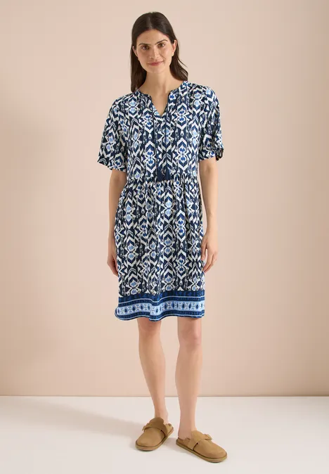 Frau trägt blau-weiß gemustertes knielanges Kleid mit kurzen Ärmeln und Taillengürtel, dazu beigefarbene Slipper.