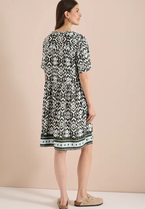Frau steht vor neutralem Hintergrund, trägt knielanges, schwarz-weiß gemustertes Kleid mit kurzen Ärmeln.