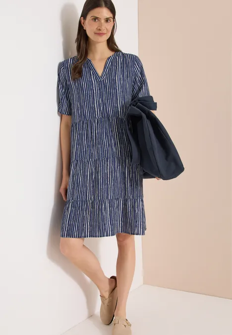 Frau trägt knieumspielendes, blau gemustertes Kleid mit V-Ausschnitt und kurzen Ärmeln, beige Schuhe, Jacke über der Schulter.