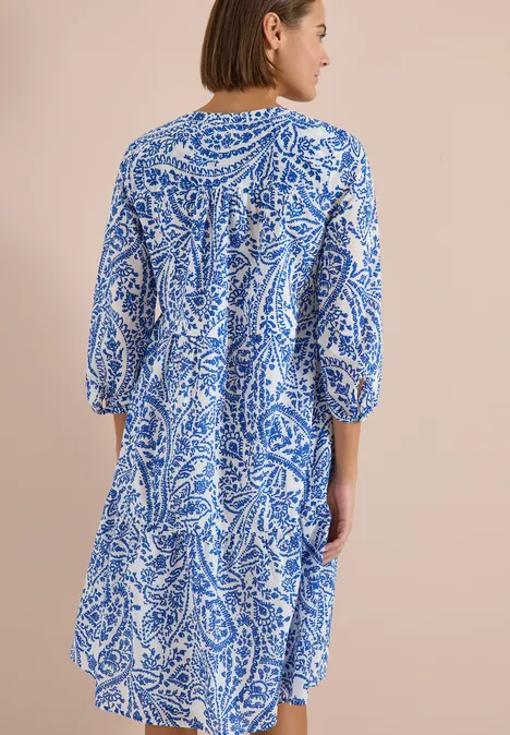 Frau von hinten in lockerem, knielangem Kleid mit blau-weißem Blumenmuster vor neutralem Hintergrund.