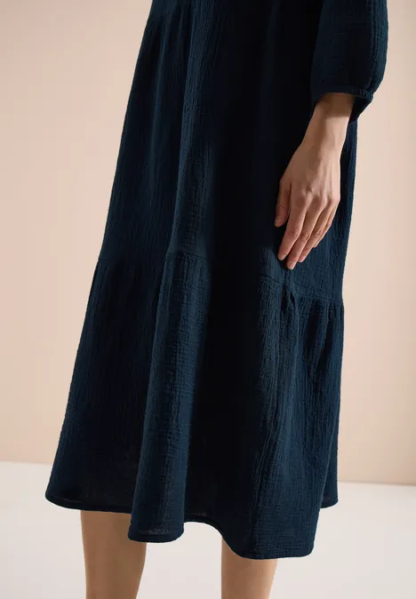 Ein dunkelblaues, knitteriges Midikleid mit weitem Rock, getragen von einer Person, Hand seitlich vor hellem Hintergrund.