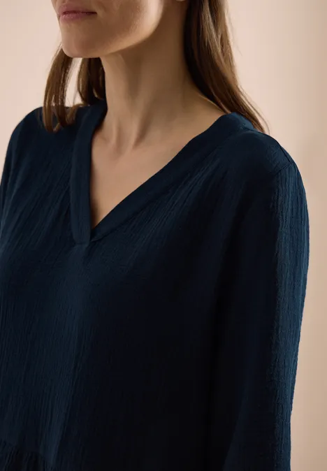 Eine Frau trägt eine dunkelblaue, strukturierte V-Ausschnitt-Bluse vor hellem Hintergrund.