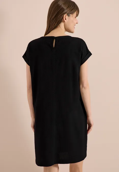 Vrouw van achteren in een zwarte, losvallende jurk met korte mouwen tegen een neutrale achtergrond.