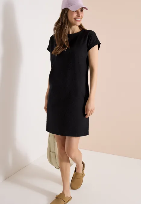 Vrouw in simpel zwart jurkje met korte mouwen, pet, beige sloffen en tas voor een lichte muur.