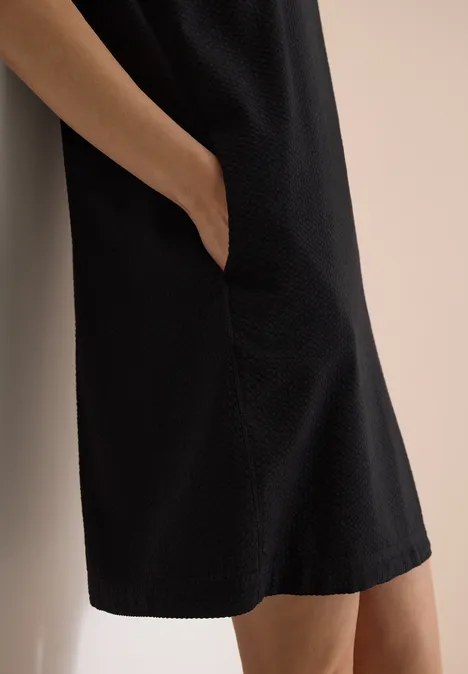 Zwarte knielange jurk met zijzak, gedragen voor een lichte, neutrale achtergrond.