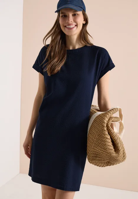 Frau in dunkelblauem T-Shirt-Kleid und Cap trägt eine geflochtene Strohtasche.