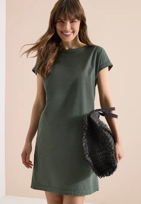 Eine Frau in einem grünen Kurzarmkleid hält eine schwarze Tasche vor hellem Hintergrund.