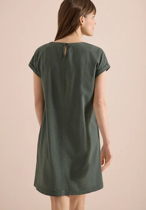 Frau mit Rücken zur Kamera in schlichtem, locker sitzendem, kurzärmligem, dunkelgrünem Kleid.