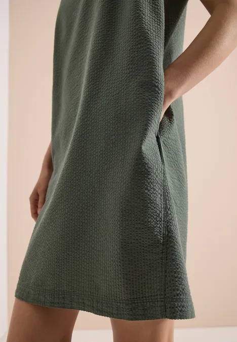 Eine Person trägt ein grün strukturiertes, knielanges Kleid mit seitlicher Tasche.