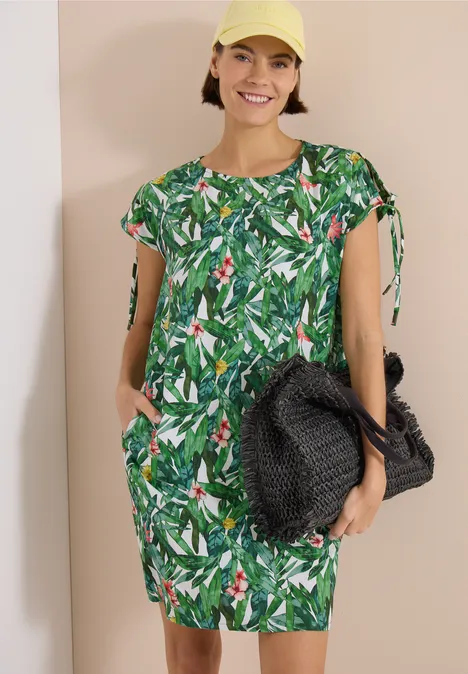 Knielanges Kleid mit floralem Muster khaki Frau in kurzärmeligem, grünem Kleid mit tropischem Blattmuster, Seitentaschen und großer schwarzer Korbtasche.