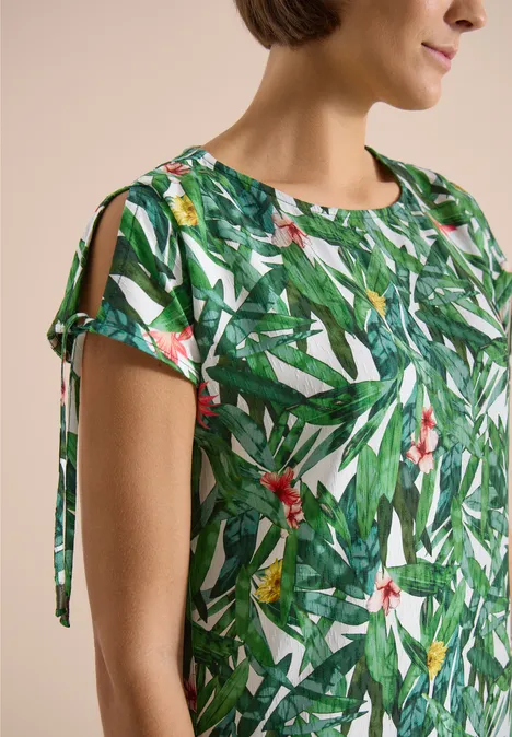 Knielanges Kleid mit floralem Muster khaki Naheaufnahme einer Frau in grüner Bluse mit tropischem Blatt- und Blumenprint und Ärmeln mit Bändchen.