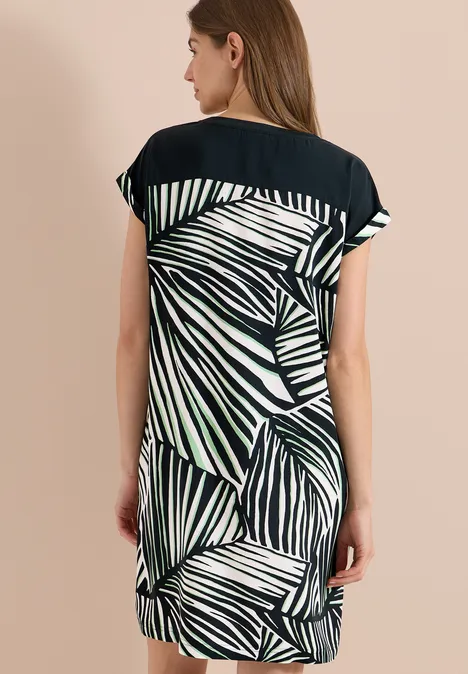 Frau von hinten in lockerem Kurzarm-Kleid mit schwarzem Oberteil und grün-weißem Blätterprint.