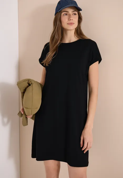 Frau in schwarzem T-Shirt-Kleid mit Kapuze hält eine grüne Tasche vor beiger Wand.