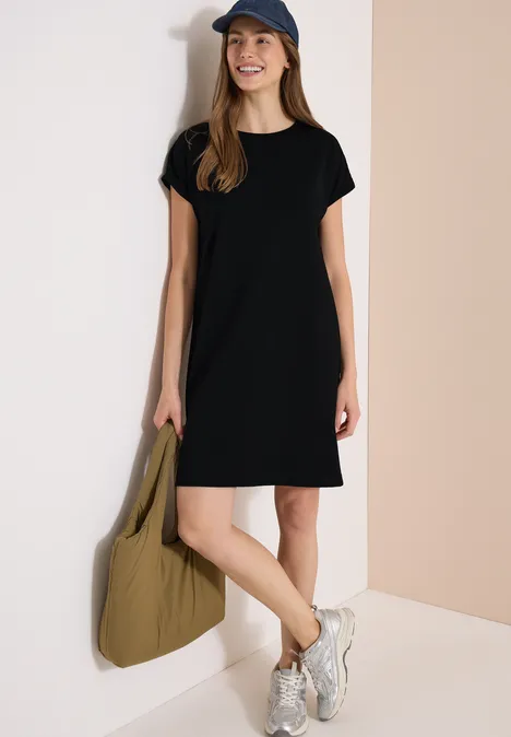 Frau in schwarzem T-Shirt-Kleid, Baseballcap, weißen Sneakers und großer olivgrüner Tasche.