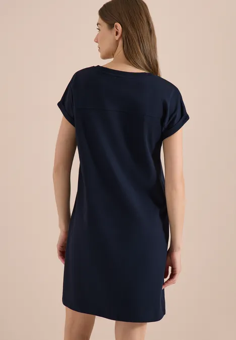 Frau von hinten in schlichtem knielangem, dunkelblauem T-Shirt-Kleid vor beigem Hintergrund