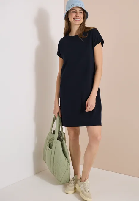Frau in lockerem dunkelblauem T-Shirt-Kleid, hellen Sneakern, grüner gesteppter Tasche und Cap.