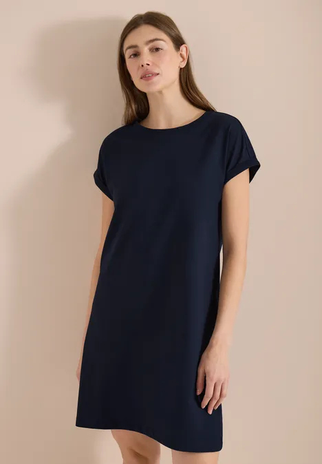 Frau in schlichtem, dunkelblauem T-Shirt-Kleid mit kurzen Ärmeln vor neutralem Hintergrund.