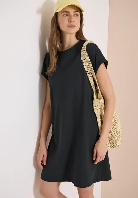Eine Frau trägt ein schwarzes T-Shirt-Kleid und hält eine geflochtene Strohtasche vor hellem Hintergrund.